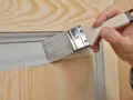 Une main utilise un pinceau pour relooker une porte avec des moulures, appliquant une peinture grise sur le bois clair.