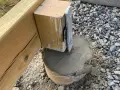 En trästolpe fästs i en betongplint, en del av arbetet med att bygga altan på plintar. Grus och jord syns runt plinten.