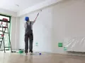 Malowanie ścian w nowym mieszkaniu. Pracownik w kasku maluje wałkiem. Ciekawe, ile za metr malowania?