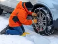 Uomo in giacca arancione monta catene da neve su ruota posteriore. Le catene da neve devono essere montate sulle ruote posteriori per la massima aderenza.