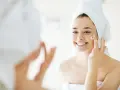 Una mujer sonriente con toalla en la cabeza se aplica crema en la cara, un paso esencial para como cuidar la piel de la cara.
