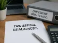 Dokumenty dotyczące zawieszenia działalności i ZUS. Na biurku leży dokument "Zawieszenie działalności", segregator z tym samym napisem, kalkulator i laptop.
