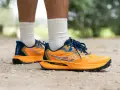 Gambe di persona che indossa scarpe da trail running Saucony arancioni e blu su un sentiero sterrato.