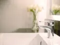 Rubinetto cromato o acciaio lucido su lavandino bianco, con fiori sfocati sullo sfondo.
