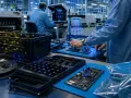 Workers in a cleanroom assemble electronics on a blue mat, surrounded by electrically safe plastics and components.