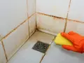 Main d'une personne portant un gant orange, utilisant une éponge jaune pour nettoyer le joint de carrelage sale dans une douche.