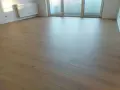 Jasne panele ułożone wzdłuż pomieszczenia, tworząc wrażenie większej przestrzeni. W tle biały grzejnik i okno balkonowe.