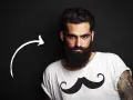 Hombre con barba tupida, quizás decidiendo dejarse barba por primera vez. Camiseta blanca con estampado de bigote.