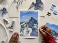 Une main peint une peinture montagne facile, avec des tubes de peinture et des nuanciers bleus à proximité.