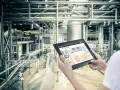 Operator kontroluje linię produkcyjną za pomocą tabletu, na którym widnieje wykres zużycia energii. W tle widać skomplikowany system rur i taśmociągów. Czujnik podczerwieni może być kluczowy dla monitorowania procesów.
