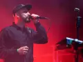 Wokalista z brodą i czapką na głowie śpiewa do mikrofonu na scenie. Czerwone światła tworzą atmosferę koncertu. Tauron Nowa Muzyka line-up.