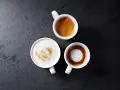 Trzy białe filiżanki z kawą: espresso, latte macchiato i kawą z pianką na ciemnym tle.