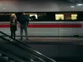 Para stojąca na peronie przed pociągiem, obok schodów ruchomych.