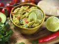 Frisches Guacamole-Rezept in einer gelben Schüssel, garniert mit Limettenscheiben. Daneben liegen Avocado, Koriander, Tomaten und Chili.