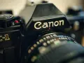 Zbliżenie na czarną lustrzankę Canon AE-1. Widoczny napis "Canon" i fragment obiektywu. Idealny do połączenia z aparatem.