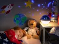 Śpiące dziecko pod kocem w gwiazdki, obok pluszowy miś w stroju astronauty. Na ścianie rakieta i Ziemia, a na suficie projektor dla dzieci na sufit wyświetla gwiazdy.