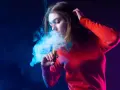 Joven exhala humo de un vaper, rodeada de luces azules y moradas.
