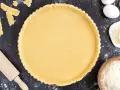 Ein vorbereiteter Mürbeteigboden in einer Tarteform, bereit für das 1 2 3 Teig Rezept. Zutaten wie Eier, Butter und Mehl sind daneben zu sehen.