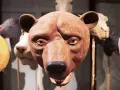 En brun björnmans ansikte, gjord av papier mache lim, med andra djurmasker i bakgrunden.