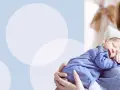 Uśmiechnięta mama tuli śpiącego noworodka w pasiastym pajacyku. Wskazówki, jak ubierać noworodka w domu.