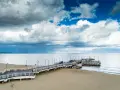 Długie molo w Brzeźnie wchodzi w spokojne morze. Na piaszczystej plaży widać kilka osób i samochody na molo.