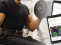 En man utför bicepscurl med stång. En surfplatta visar övningar för armar, inklusive "Spider Curls".