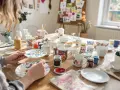 En person målar blommor på en mugg. Bordet är fullt av muggar, tallrikar och färger, redo för kreativt måla på muggar.
