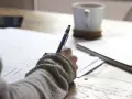 Ręka pisze długopisem na papierze, planując, jak napisać książkę. Obok kubek kawy i notatnik.