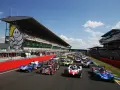 Una fila de coches de carreras listos para comenzar las 24 horas de Le Mans. El icónico muñeco de Michelin preside la tribuna.
