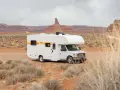 En vit husbil med "Indie Campers"-logotypen står på en grusväg i ett ökenlandskap med röda klippformationer. En perfekt start för att besöka Jylland.