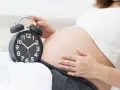 Zbliża się termin porodu. Kobieta trzyma dłoń na brzuchu obok budzika.