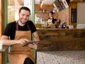 Hombre sonriente con delantal usa tablet en su cafetería, gestionando pedidos con un sistema erp restaurantes.