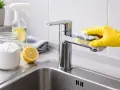 Rękawiczka w żółtej rękawiczce czyści kran gąbką, pokazując, jak wyczyścić sitko w kranie naturalnymi środkami: sodą i cytryną.