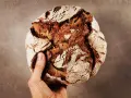 Hand hält ein rustikales Brot aus Emmer Urkorn mit knuspriger Kruste und dunkler Krume.