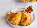 Tres empanadillas al horno doradas y crujientes, servidas en un plato blanco con un pequeño cuenco de salsa de tomate y una hoja de perejil.