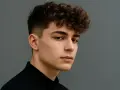 Młody mężczyzna z kręconymi, puszystymi włosami (fryzura fluffy hair) i wygolonymi bokami, ubrany w czarną koszulę, na szarym tle.