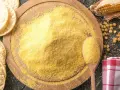 Stos mąki kukurydzianej na desce, obok łyżka z mąką, kukurydza i wafle.