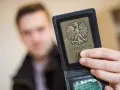 Komornik pokazuje legitymację z orłem. W tle niewyraźna postać mężczyzny, być może w związku z długiem osób prywatnych.