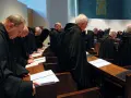 A group of Italian monks in black robes stand in a church, heads bowed over open prayer books.