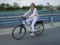 Kobieta w białych spodniach i sweterku jedzie na rowerze elektrycznym. W tle widać most i budynki.