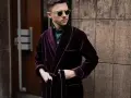 Elegancki mężczyzna w bordowej bonżurce z aksamitu, w okularach przeciwsłonecznych, stoi przy kamiennej ścianie.