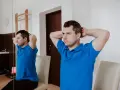 Mężczyzna w niebieskiej koszulce wykonuje ćwiczenia na szyję, opierając dłonie o tył głowy.