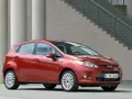 Czerwony Ford Fiesta MK6 zaparkowany przed nowoczesnym budynkiem z betonowymi kolumnami i schodami.