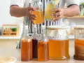 Personne tenant une bouteille et un verre de kombucha, entouré de plusieurs bouteilles et bocaux contenant le **champignon de kombucha** en fermentation.