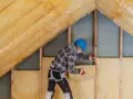 Pracownik w kasku montuje wełnę mineralną, wykonując ocieplanie domu na poddaszu.