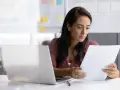 Kobieta w biurze czyta dokumenty obok laptopa.