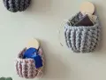 Three crocheted wall pockets in grey and pink, one holding a wallet, another blue ribbon, on a light wall.