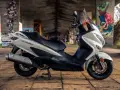 Biały skuter Suzuki Burgman 125, z czarnym siedzeniem, zaparkowany pod mostem z kolorowymi graffiti. Idealny do miasta, sprawdź suzuki burgman 125 dane techniczne.
