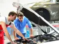 Zwei Männer blicken in den offenen Motorraum eines Autos. Der Mechaniker in Rot erklärt dem Kunden, welche Werkstatt mitgebrachte Teile einbaut.