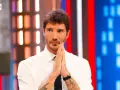 Santiago de Martino, con le mani giunte in preghiera, guarda intensamente verso l'obiettivo, con uno sfondo di luci colorate.